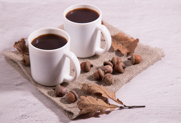 Deux tasses blanches de café de gland sur une serviette avec des glands et des feuilles de chêne sur fond clair