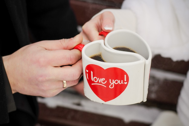 Photo deux tasses blanches en forme de cœur entre les mains de la mariée et du marié. saint valentin, le jour de l'amour et du mariage