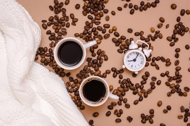 Deux tasses de café noir, réveil, pull blanc et grains de café sur fond beige. Rendez-vous cosy et bien-être. Vue de dessus