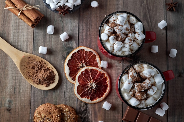 Photo deux tasses rouges de chocolat chaud avec de la guimauve saupoudrée de poudre de cacao sur une table en bois avec des tranches de pamplemousse séché et des morceaux de chocolat. boisson chaude d'hiver aux épices. vue de dessus.