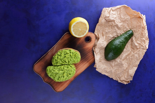 Deux toasts d'avocat vert frais sur une planche de bois Toasts un demi-avocat et un citron jaune dans une peau sur un fond bleu Gros plan