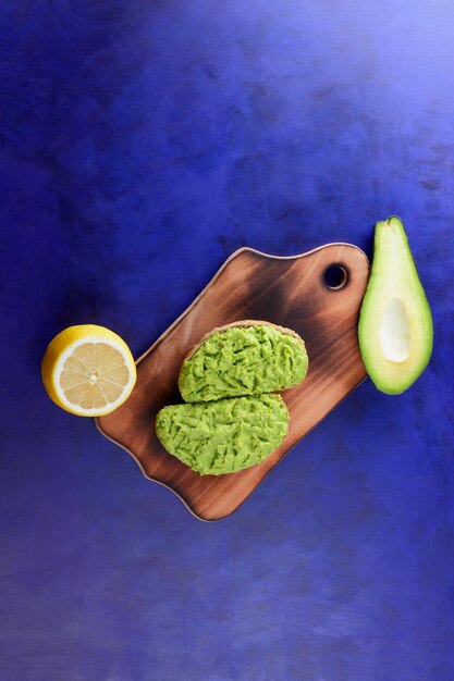 Deux toasts d'avocat vert frais sur une planche de bois Toasts un demi-avocat et un citron jaune dans une peau sur un fond bleu Gros plan