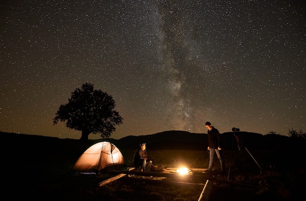 Deux touristes au feu de camp en feu devant la tente, appareil photo sur trépied sous un ciel étoilé sombre.