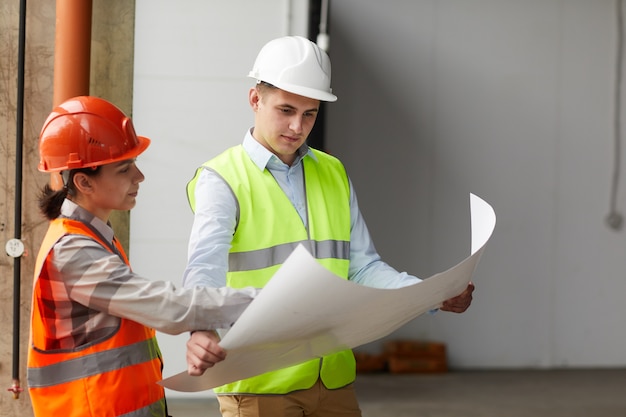 Deux travailleurs de la construction dans des casques de travail examinant le plan ensemble en se tenant debout dans un bâtiment vide