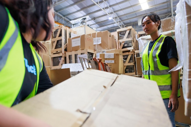 Deux travailleuses transportant et soulevant une lourde boîte en carton Ouvrière d'entrepôt obèse asiatique en gilet de sécurité et casque de sécurité travaillant dans l'usine d'entrepôt industrielle