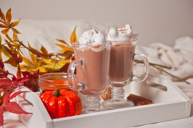 Deux verre de cacao crémeux chaud avec de la mousse sur le plateau blanc avec des feuilles d'automne et des citrouilles sur le fond