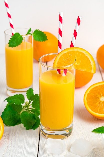 Deux verres de jus d'orange avec des tubes, de la menthe et des oranges fraîches sur un bureau blanc.