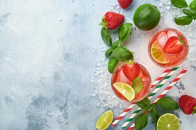 Deux verres de punch et des ingrédients frais pour faire de la limonade, de l'eau détox infusée ou un cocktail. Fraises, citron vert, menthe, basilic, glaçons et shaker sur fond gris pierre ou béton. Vue de dessus.