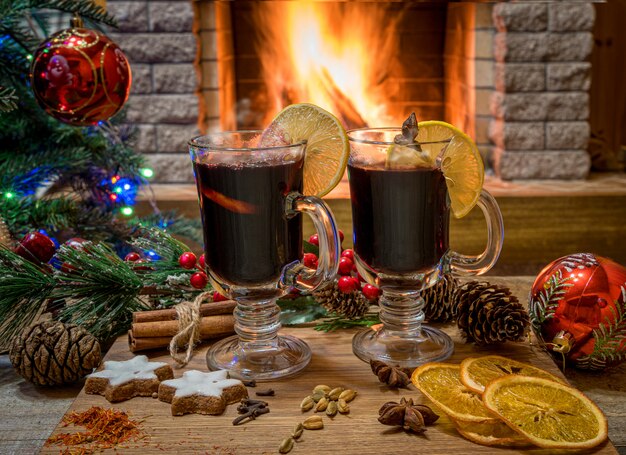 Deux verres de vin chaud sur une planche de bois avant l'arbre de Noël décoré de jouets et de lumières de Noël en face de la cheminée en feu.