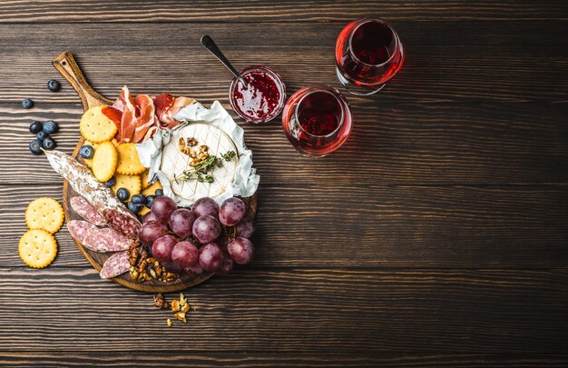 Deux verres de vin rouge, planche de bois, charcuterie, fromage, fruits, trempette. Fond en bois