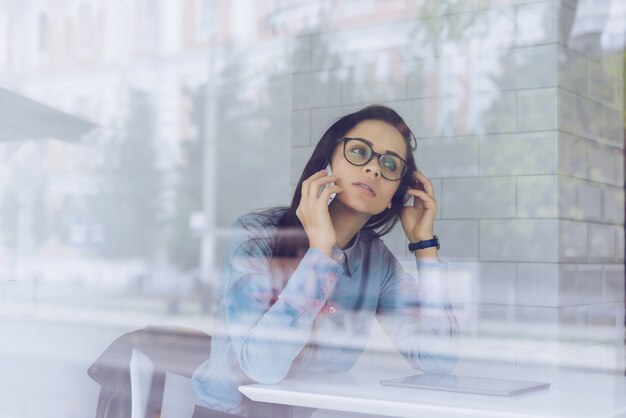 Devant la fenêtre, une femme est assise à une table et parle au téléphone