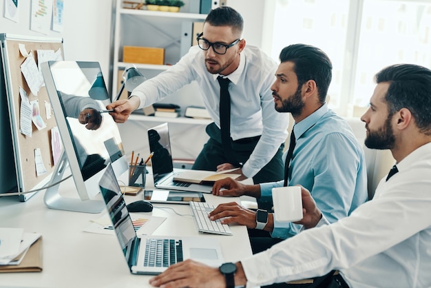 Développer de nouvelles approches. Groupe de jeunes hommes modernes en tenue de soirée travaillant à l'aide d'ordinateurs assis au bureau