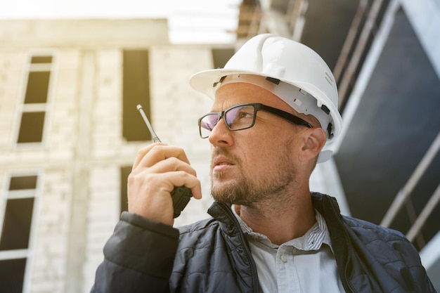 Développeur ou ingénieur masculin portant un casque de sécurité blanc à l'aide d'un talkie-walkie lors de l'inspection du chantier de construction