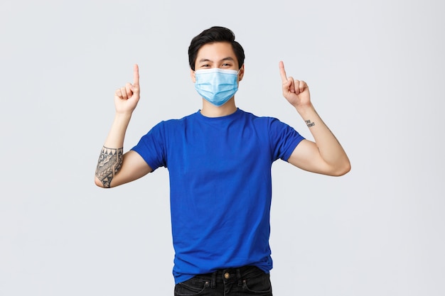 Différentes émotions, distanciation sociale, auto-quarantaine sur le coronavirus et concept de style de vie. Gai homme asiatique souriant en masque médical et t-shirt, pointant les doigts vers le haut pour faire de la publicité, montrant la bannière