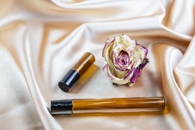 Différents récipients pour stocker les produits cosmétiques en verre foncé reposent sur les plis du tissu en satin de soie, entourés de fleurs roses séchées. Parfum produit de beauté cosmétique.