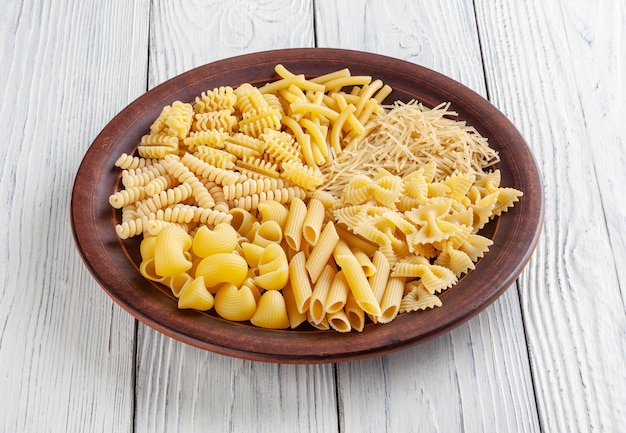 Différents types et formes de pâtes italiennes dans une assiette en céramique sur fond de bois blanc