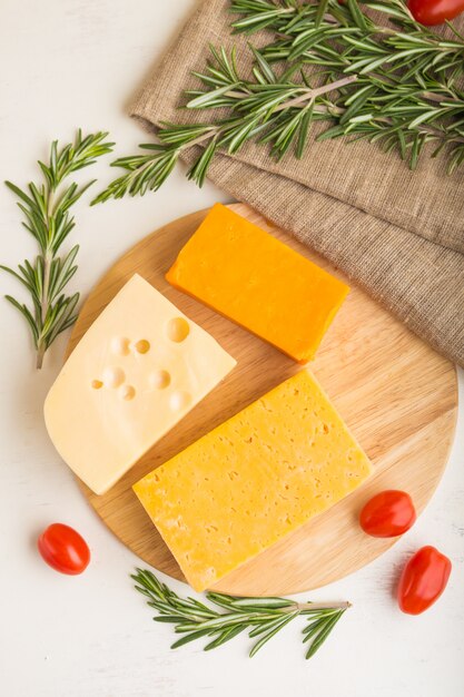 Différents types de fromage au romarin et tomates sur planche de bois sur une surface en bois blanche. Vue de dessus, gros plan.