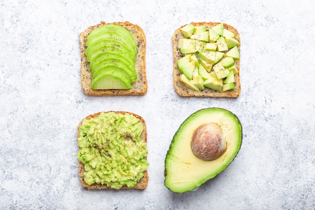 Différents types de toasts à l'avocat et demi d'avocat entier sur fond de pierre blanche, concept. Façons de préparer et de servir un délicieux sandwich à l'avocat pour le petit-déjeuner sain, vue de dessus, gros plan