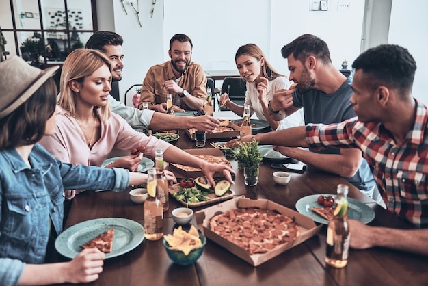 Le dîner est sur la table. Groupe de jeunes en tenue décontractée mangeant et souriant tout en organisant un dîner