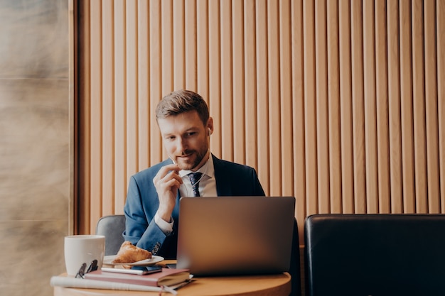 Directeur de bureau masculin portant un costume bleu et une chemise blanche, parlant à ses employés lors d'une réunion en ligne et regardant un ordinateur portable assis et travaillant à distance dans un café pendant un voyage d'affaires