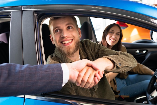 Le directeur des ventes serre la main d'un homme qui est assis dans une nouvelle voiture Un couple marié a acheté sa propre voiture