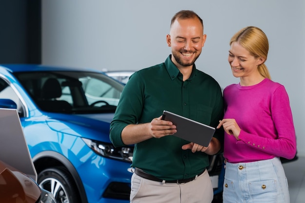 Photo un directeur des ventes souriant offre à une jeune femme des options pour acheter une nouvelle voiture sur une tablette dans le contexte de la salle d'exposition