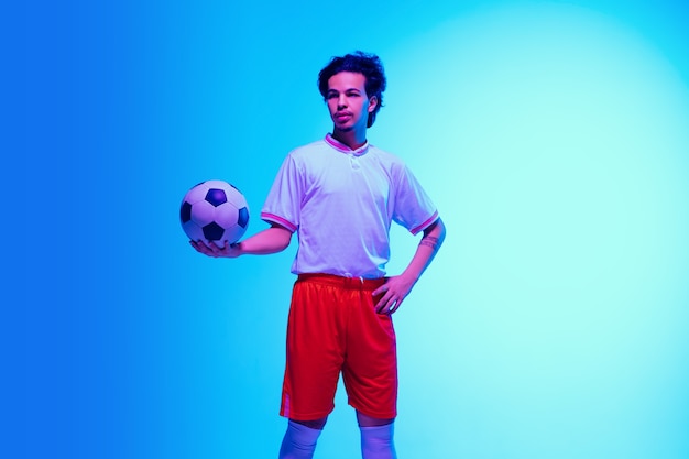 Dirigeant. Joueur de football ou de football sur un mur de studio bleu dégradé à la lumière du néon - posant confiant avec le ballon. Espace de copie.