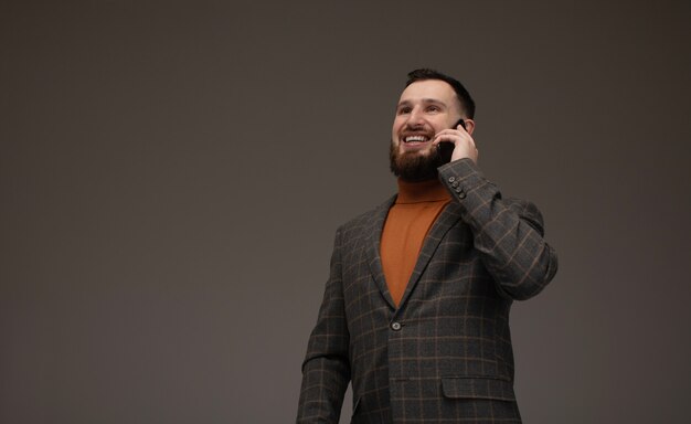 Discussion d'affaires. Souriant jeune homme barbu en costume à l'aide de téléphone intelligent. Isolé sur gris.