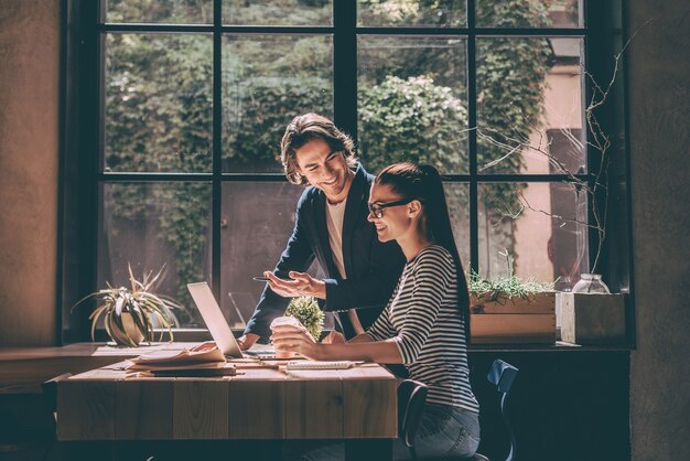 Discuter affaires ensemble. Jeune homme et femme confiants regardant un ordinateur portable et souriant tout en étant assis au bureau en bois dans un bureau ou un café créatif