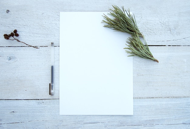 Une disposition de bureau avec une feuille de papier vide et des branches de pin sur une table en bois blanc battue