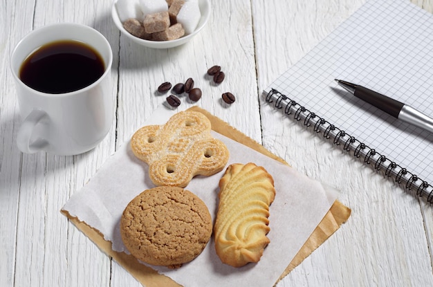 Divers biscuits, tasse de café et cahier avec stylo sur table en bois blanc