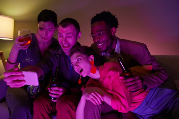 Divers groupes d'amis prenant un selfie ou une conversation vidéo ensemble lors d'une fête à l'intérieur éclairée par des lumières violettes