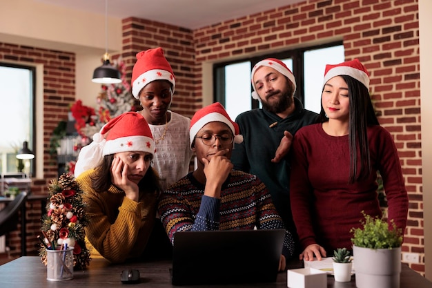 Diverses personnes festives remuant des idées au travail, portant un bonnet de noel au bureau avec un sapin de noël et des lumières. Groupe de collègues travaillant sur un projet pendant la saison des vacances d'hiver, décor de Noël.