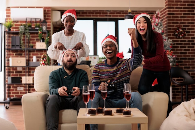 Diverses Personnes Jouant à Des Jeux Vidéo Lors D'une Fête De Noël Dans Un Bureau Festif Avec Arbre De Noël Et Ornements. Amusez-vous Avec La Compétition En Ligne Sur La Console De Télévision, Célébrant Les Vacances D'hiver.