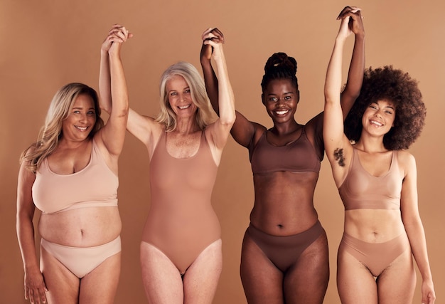 Diversité de beauté et naturel avec des amies en studio sur un fond beige avec leurs mains levées en célébration Sous-vêtements de bien-être et réel avec un groupe de femmes modèles debout ensemble