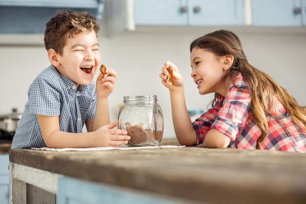 Divertissant. Adorable petit frère et soeur aux cheveux noirs exubérants riant et mangeant des biscuits assis à la table