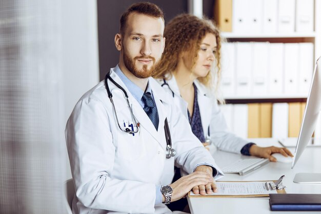 Docteur barbu avec une collègue discutant du traitement actuel de la maladie tout en étant assis sur son lieu de travail dans une clinique ensoleillée. Travail d'équipe en médecine.