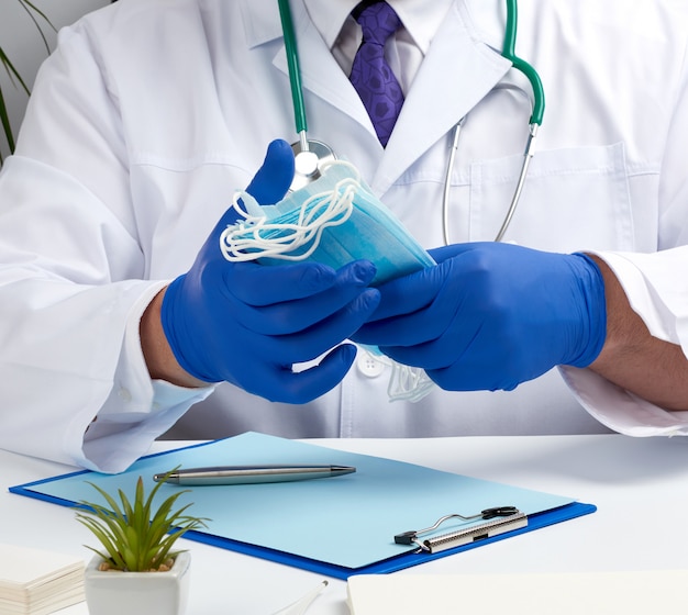 Docteur en blouse blanche et gants en latex bleu est assis à un bureau blanc et montre une pile de masques médicaux jetables