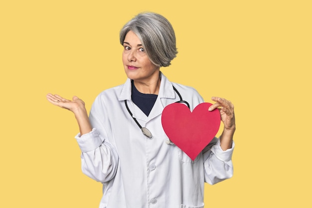 Docteur caucasien d'âge moyen avec un symbole de cœur montrant un espace de copie sur une paume et tenant une autre main sur la taille