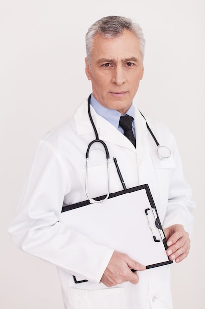 Docteur confiant. Médecin senior aux cheveux gris en uniforme regardant la caméra et tenant un presse-papiers dans ses mains tout en se tenant isolé sur blanc
