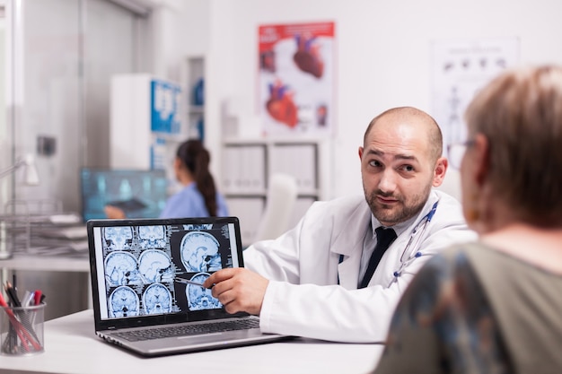 Docteur discutant avec une femme âgée avec alzheimer dans un bureau de l'hôpital pointant sur un écran d'ordinateur portable avec un scanner. Medic portant une blouse blanche. Infirmière en uniforme bleu travaillant sur ordinateur.