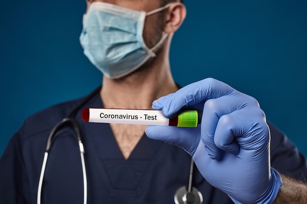 Docteur en masque, gants jetables bleus, combinaison médicale, stéthoscope sur le cou. Tenant le tube à essai de laboratoire rouge à la main. Posant sur fond bleu. Recherche sur le coronavirus. COVID-19 [FEMININE. Gros plan, espace de copie