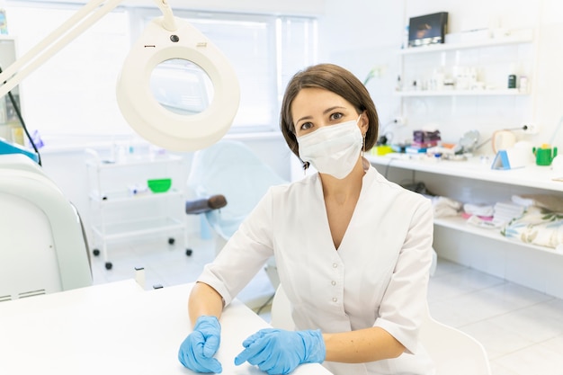 Un docteur mignon dans un manteau blanc le cosmétologue est assis dans le bureau et sourit une loupe