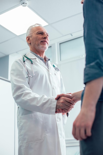 Docteur mûr gai avec un stéthoscope sur le cou souriant et serrant la main d'un patient