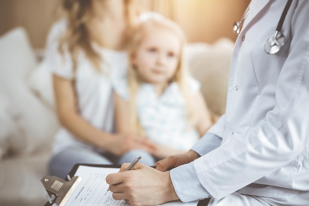 Docteur et patient. Le pédiatre utilise le bloc-notes en examinant une petite fille avec sa mère à la maison. Enfant malade et malheureux à l'examen médical.