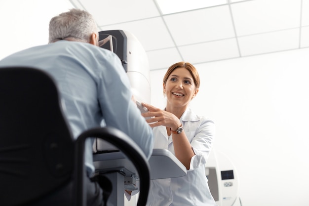 Docteur souriant. Belle jeune ophtalmologiste portant une blouse blanche souriant tout en examinant un homme à la retraite