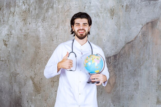 Docteur avec un stéthoscope tenant un globe terrestre et montrant un signe de main réussi signifiant un service mondial