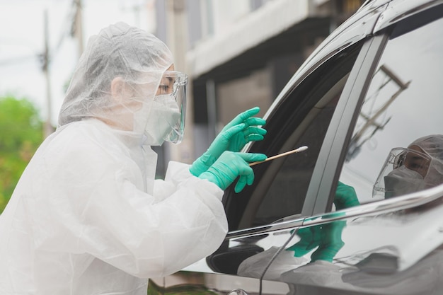Docteur en tenue de protection EPI tenant un kit de test. Travailleur médical effectuant un test Covid-19 au volant, prélevant un échantillon d'écouvillon nasal à travers la vitre de la voiture, diagnostic PCR, kit de test rapide d'antigène (ATK)