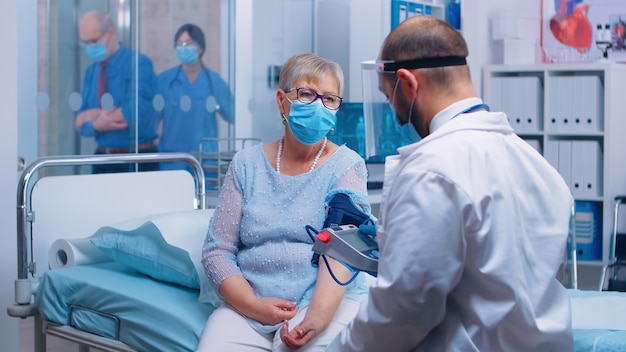 Docteur en vêtements de protection pendant COVID-19 mesurant la tension artérielle d'une vieille dame à la retraite dans un hôpital ou une clinique privé. Infirmière travaillant en arrière-plan, examinant les patients, les soins médicaux et le système de santé