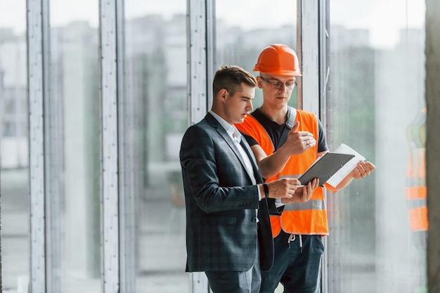 Avec des documents Un homme en costume et un bricoleur en vêtements de protection orange travaillent sur la construction